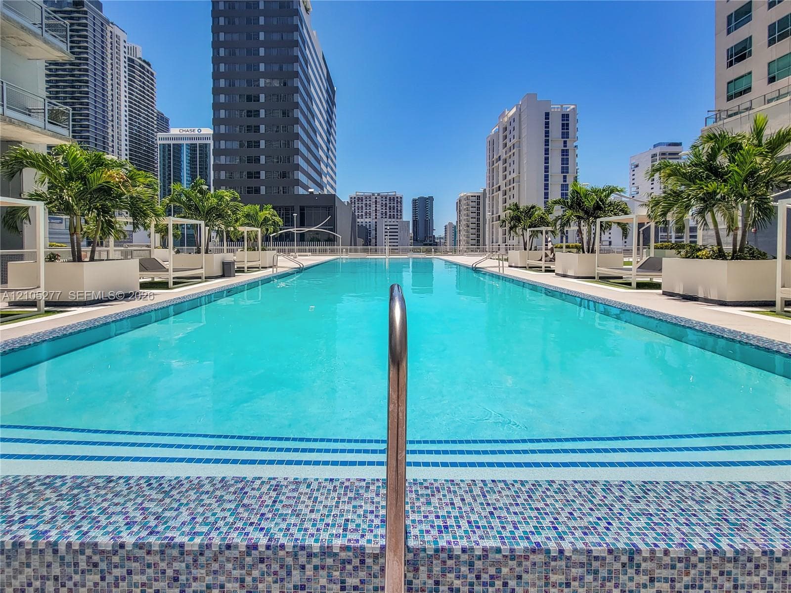 Rooftop swimming pool featuring mosaic tile detailing, surrounding palm trees in planters, and multiple cabanas