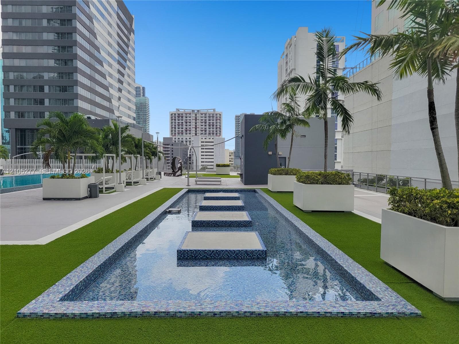 Rooftop amenity space featuring a decorative water feature with stepping stones, artificial turf, and modern planters with tropical foliage