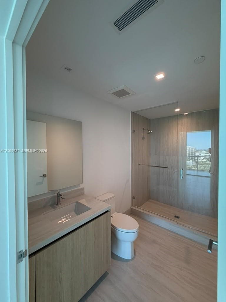 Bathroom featuring a floating vanity with a light wood-finish cabinet, integrated countertop and sink, glass-enclosed walk-in shower with large-format tile surround, and wood-finish flooring
