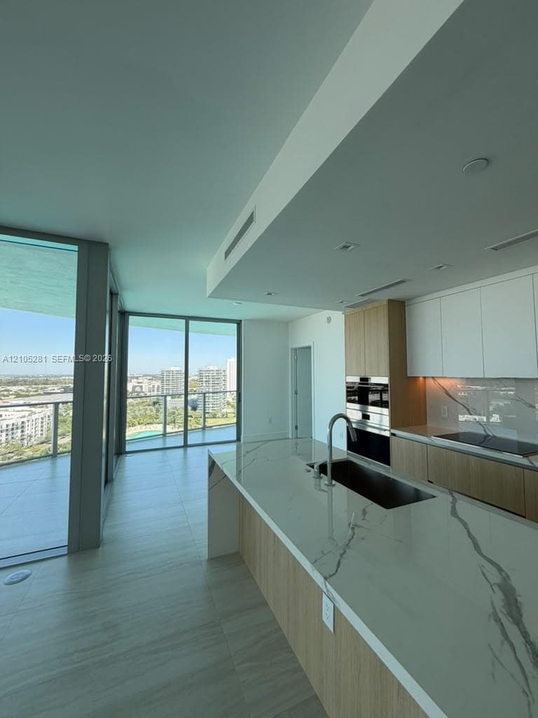 Kitchen featuring a waterfall-edge island with stone-finish countertops, flat-panel cabinetry, built-in stainless steel appliances, and floor-to-ceiling glass wall panels