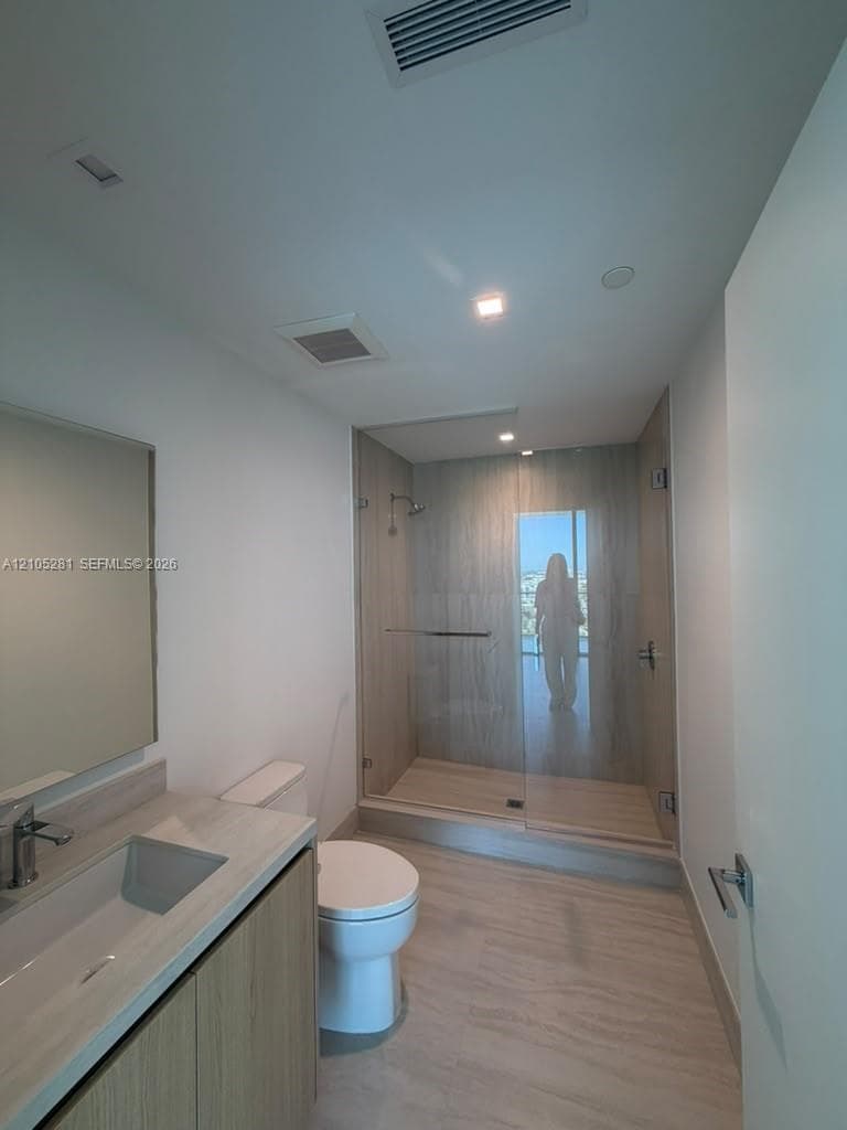 Bathroom featuring a wood-finish vanity with a neutral stone countertop, frameless glass shower enclosure with floor-to-ceiling tile, and integrated recessed lighting