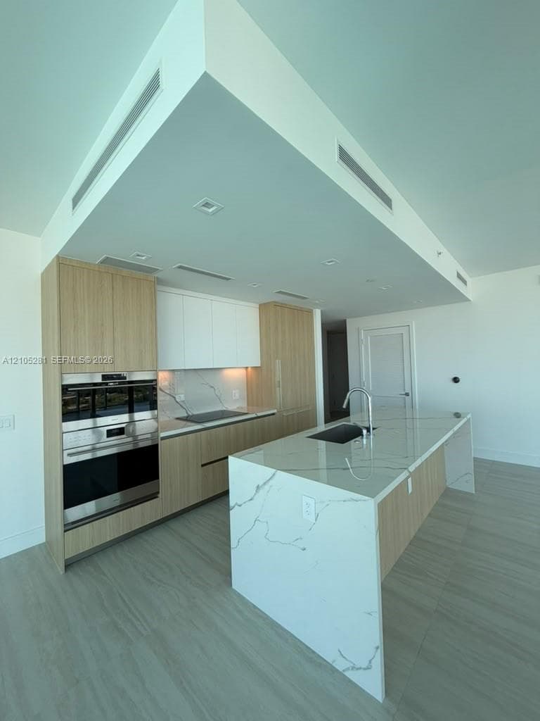 Modern kitchen featuring a waterfall-edge island, composite stone countertops, flat-panel wood-finish cabinetry, stainless steel wall-mounted oven and microwave, and wood-finish flooring