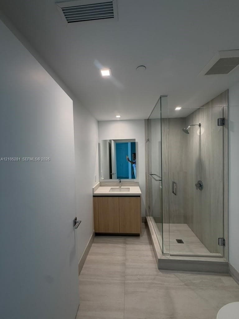 Bathroom featuring wood-finish cabinetry, a rectangular undermount sink, a glass-enclosed shower with full-height tile surround, and large-format floor tiling