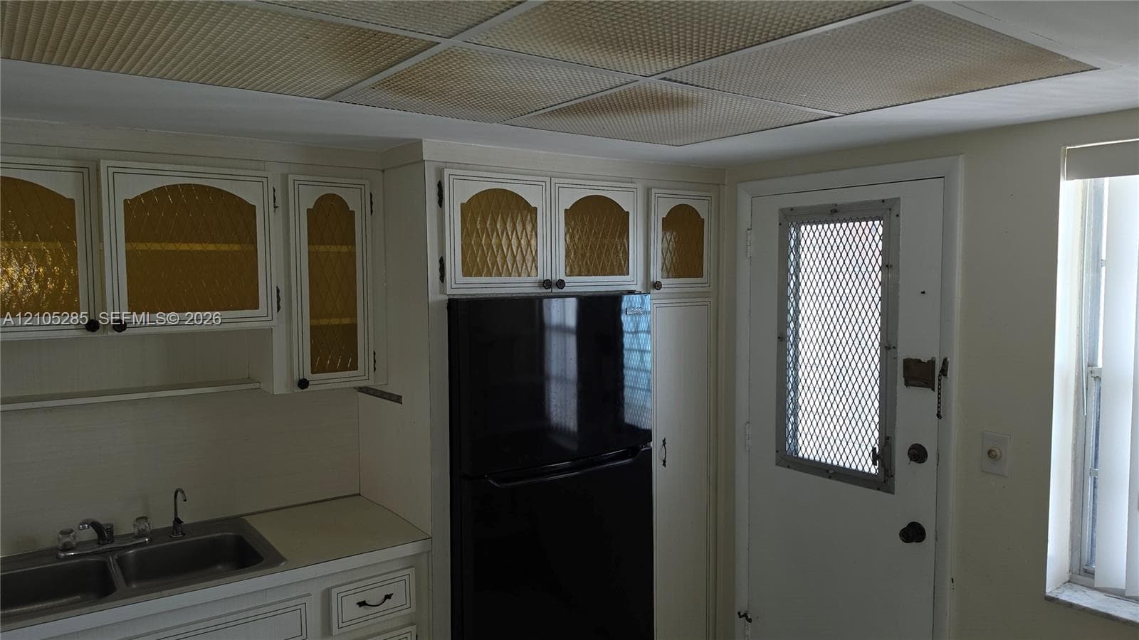 Kitchen featuring white cabinetry with textured glass inserts, double-basin stainless steel sink, black refrigerator, and drop-ceiling lighting