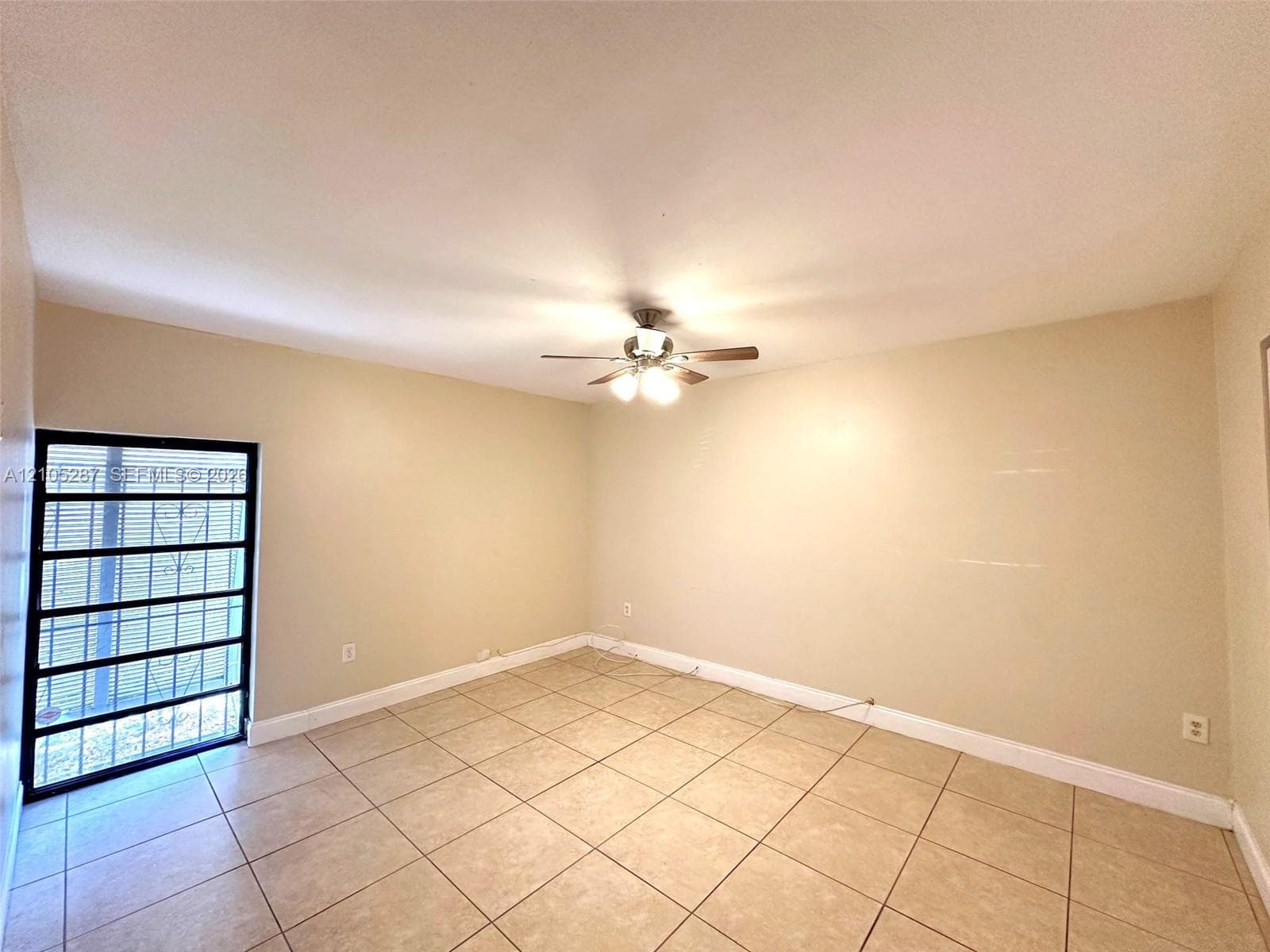 Spacious room featuring neutral-toned walls, tile flooring, a window with security bars, and a ceiling fan with integrated lighting