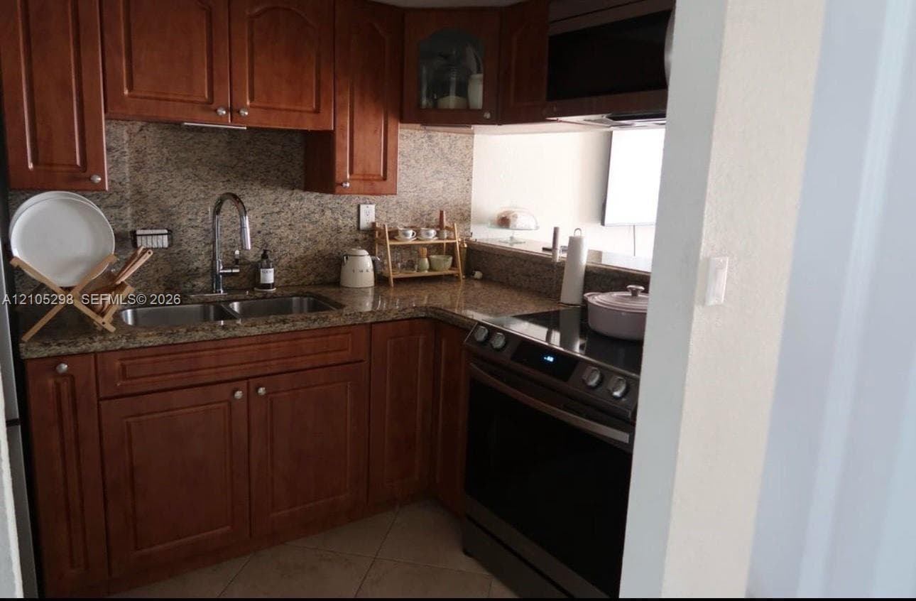 Wood-finish cabinetry with stainless steel hardware, integrated stainless steel sink, stone countertops and backsplash, and a built-in microwave