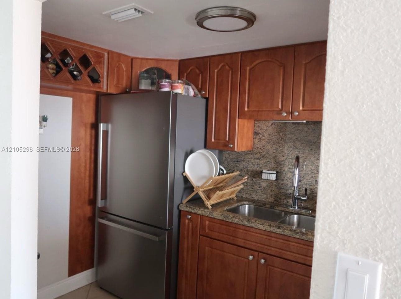 Kitchen featuring wood cabinetry, stainless steel refrigerator, double basin sink with a gooseneck faucet, and stone-finish countertops with a matching backsplash