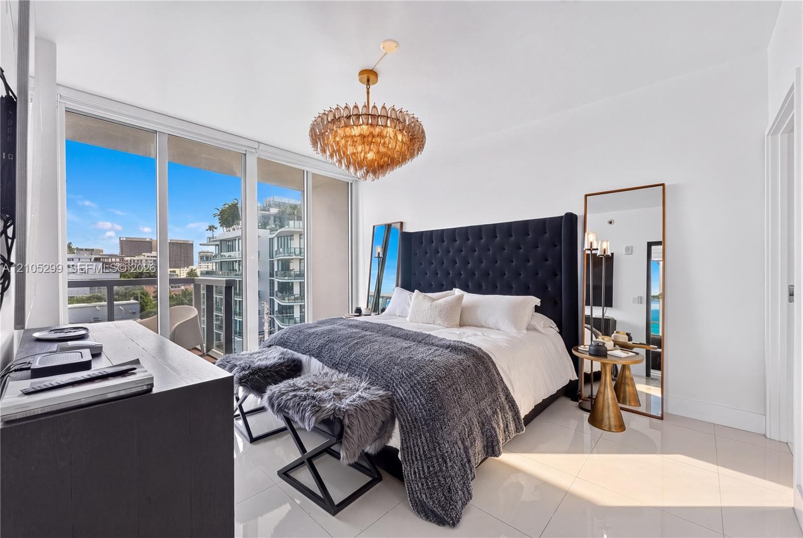 Floor-to-ceiling glass sliding doors, white tile flooring, gold-toned chandelier, dark upholstered headboard, and a built-in dresser