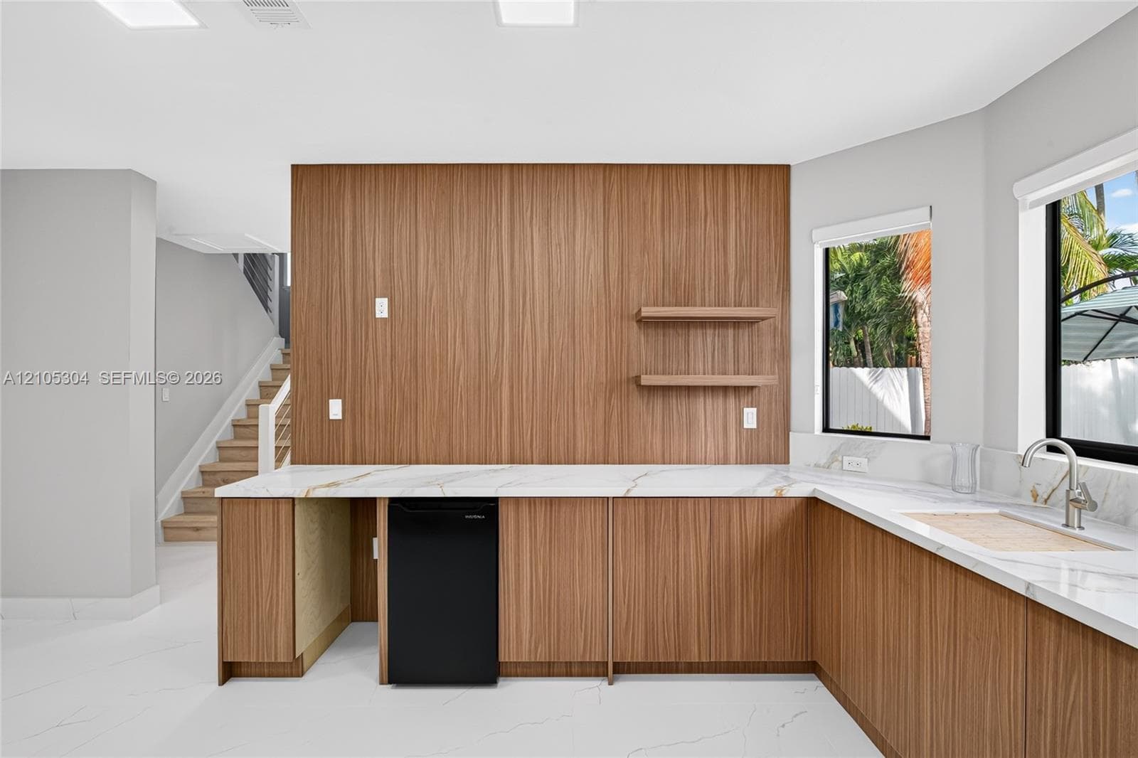 Custom wet bar featuring wood-veneer cabinetry, stone-finish countertops, floating shelving, an undermount sink with a gooseneck faucet, and integrated beverage refrigeration