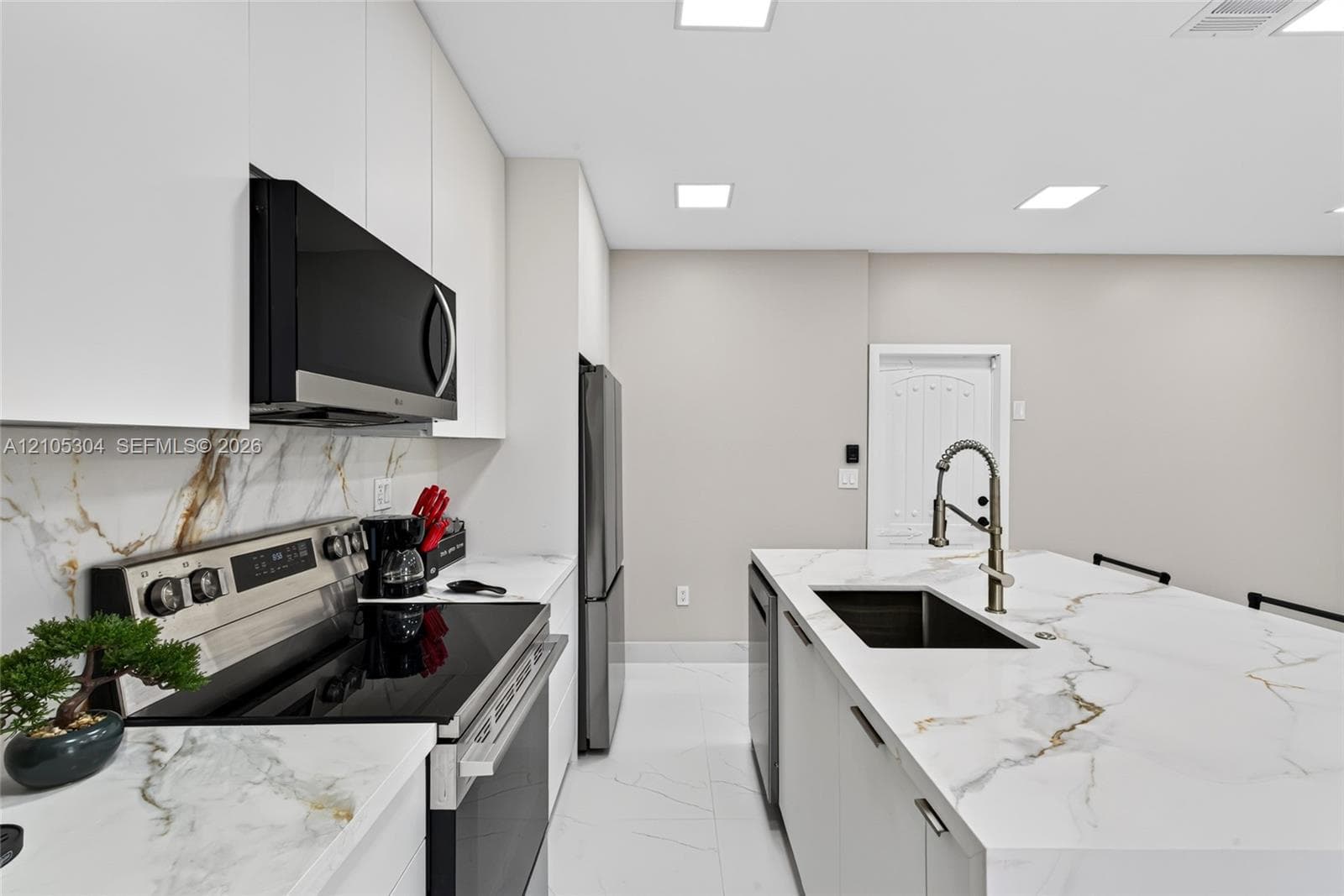 Kitchen featuring white slab cabinetry, large-format tiled flooring, stone-finish countertops with matching full-height backsplash, stainless steel appliances, and a central island with a high-arc faucet