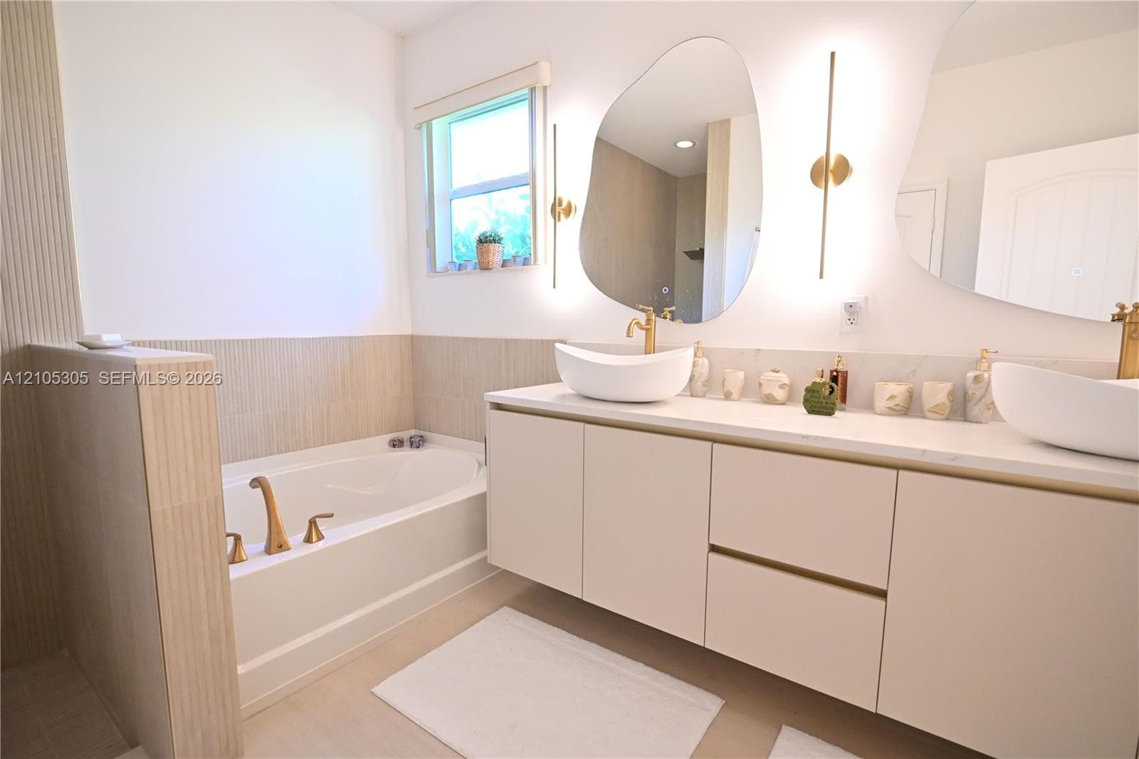 Built-in soaking tub with gold-tone fixtures, dual floating vanities with vessel sinks, stone countertops, and back-lit wall-mounted mirrors