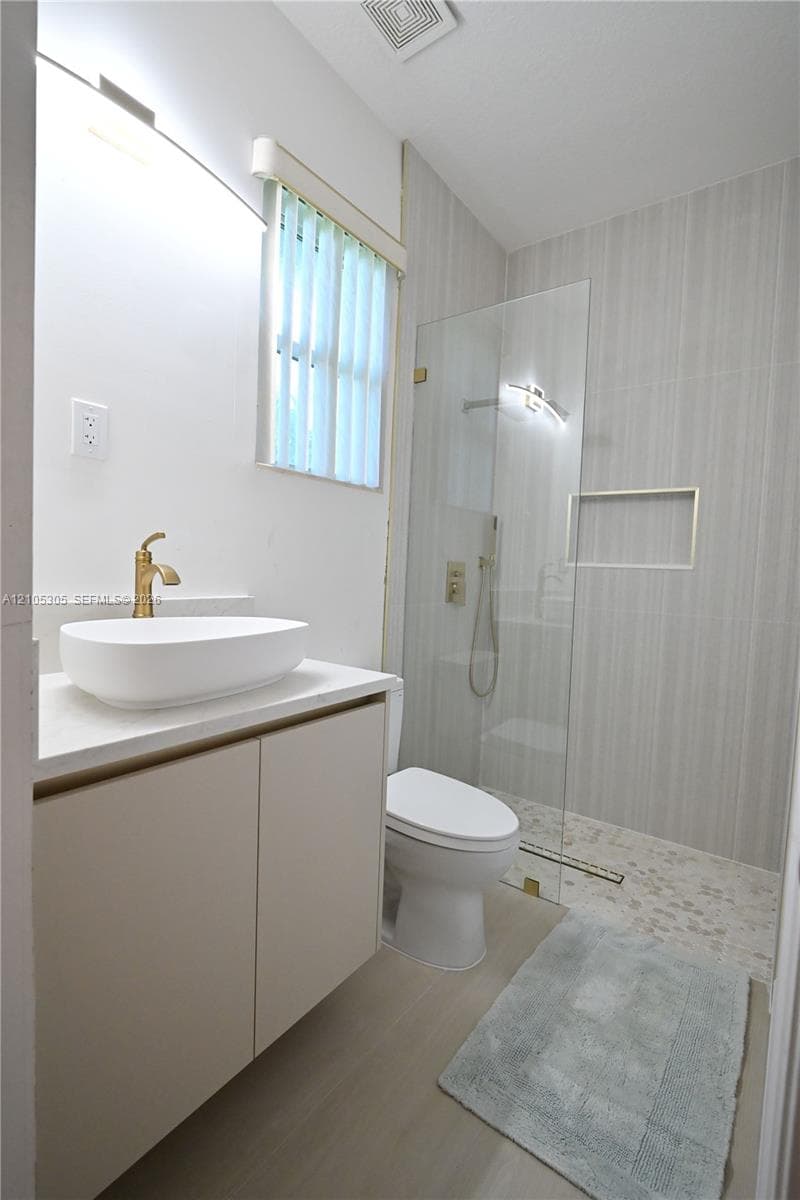 Floating vanity with a vessel sink and gold-tone faucet, glass-enclosed shower with a tiled wall and pebble floor, and wood-finish flooring