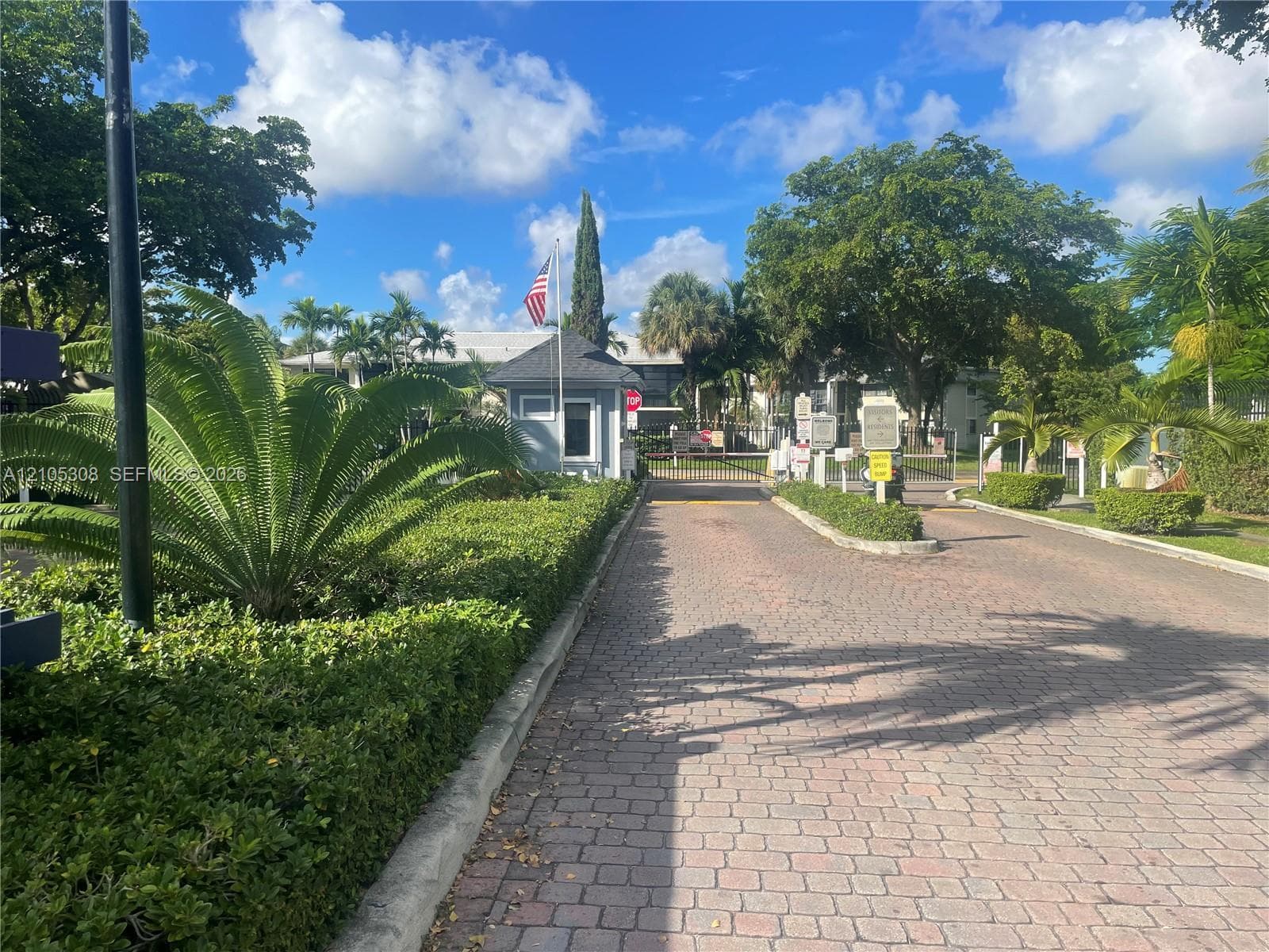 Gated entrance featuring a brick-paver driveway, security guardhouse, and mature landscaping