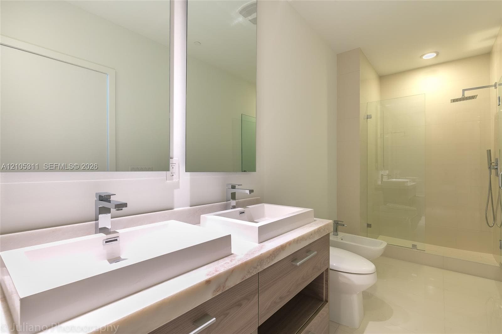 Dual rectangular vessel sinks with chrome faucets, wood-finish vanity, large wall-mounted mirror with integrated lighting, glass-enclosed shower with rainfall showerhead, and a bidet
