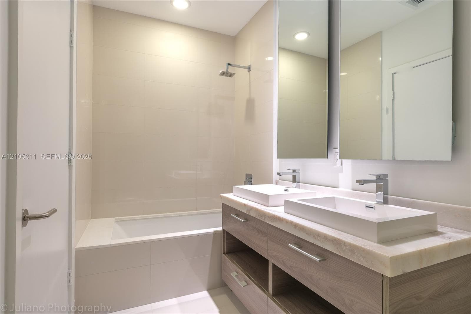 Built-in bathtub with tiled surround, dual rectangular vessel sinks, wood-finish vanity with open shelving, and wall-mounted medicine cabinets