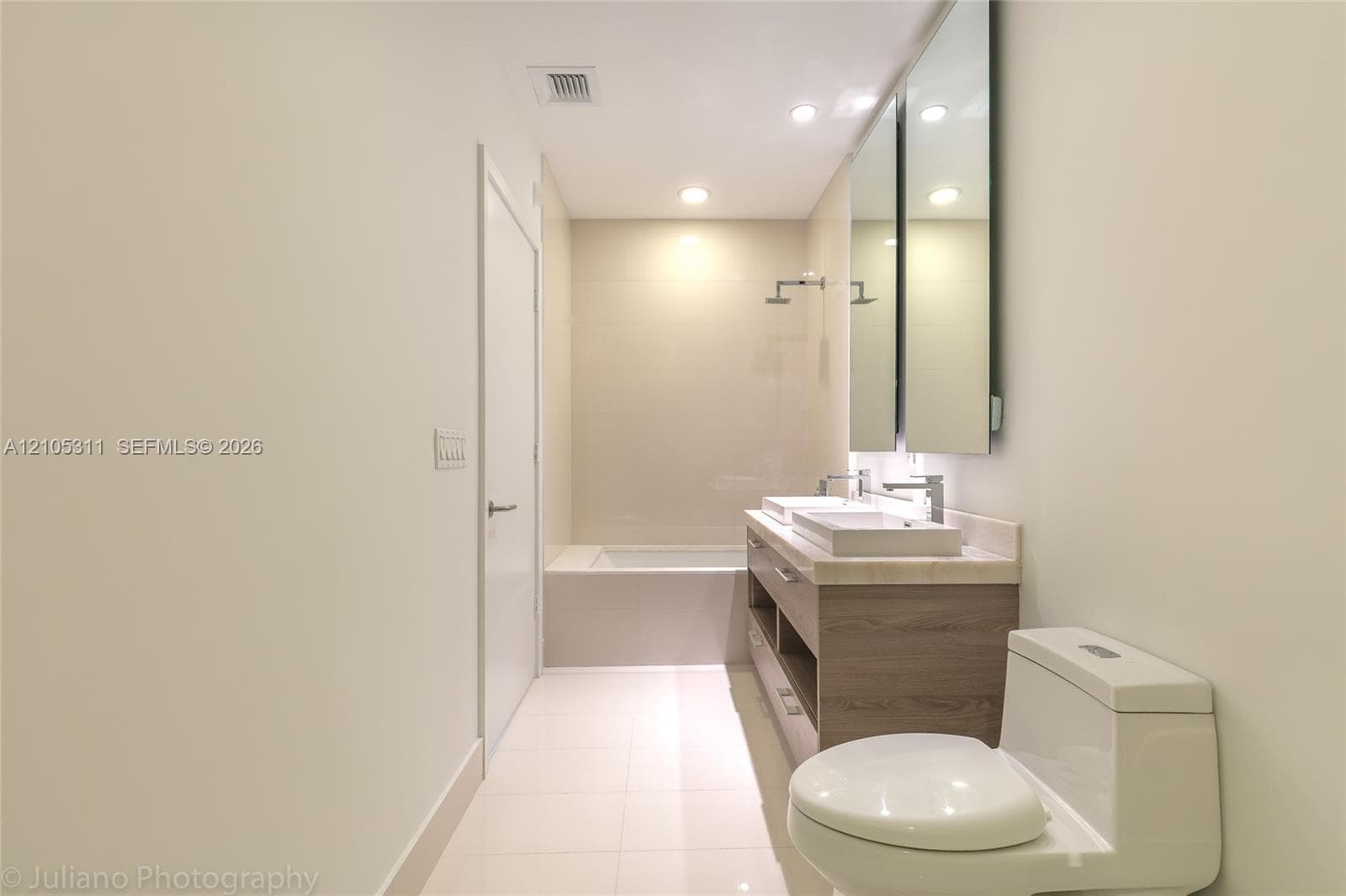 Dual vessel sinks on a vanity with wood-finish cabinetry, a built-in bathtub, and recessed lighting