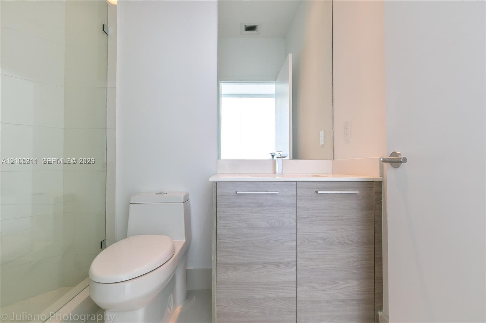 Glass-enclosed shower with white tiled walls, wood-finish vanity with a light countertop, and a wall-mounted mirror