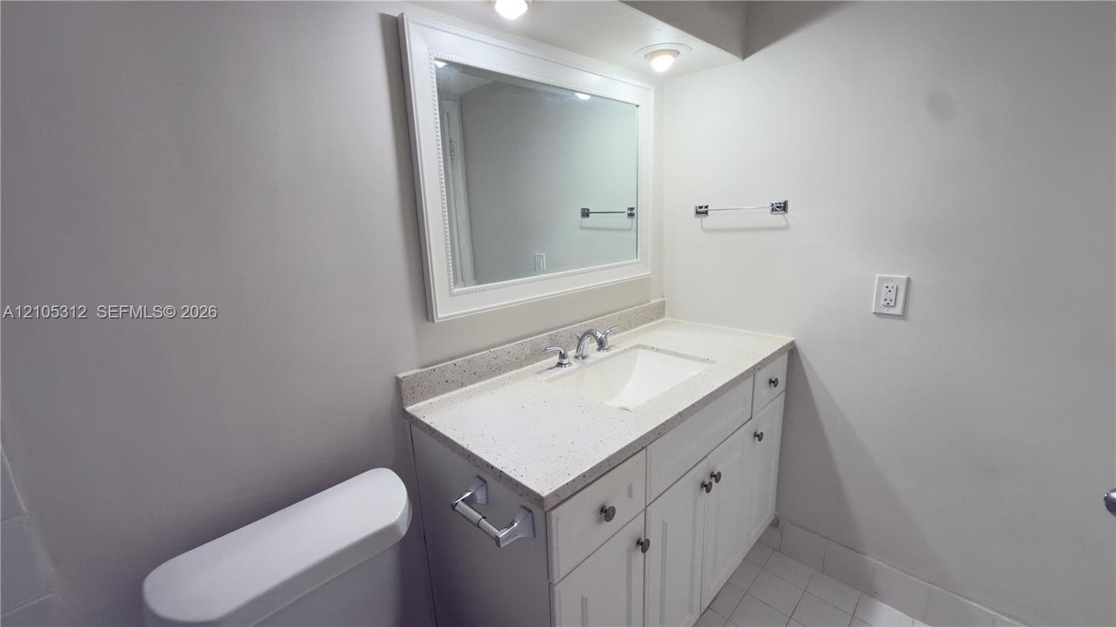 White vanity with stone-composite countertop and integrated sink, framed wall mirror, recessed lighting, and tile flooring