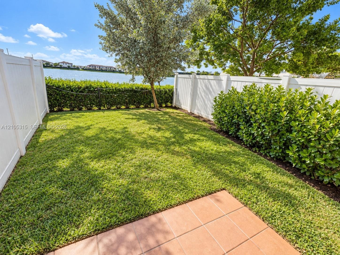 Manicured lawn with mature trees and shrubs, bordered by a white privacy fence and a hedge, featuring a tiled patio area and a visible body of water