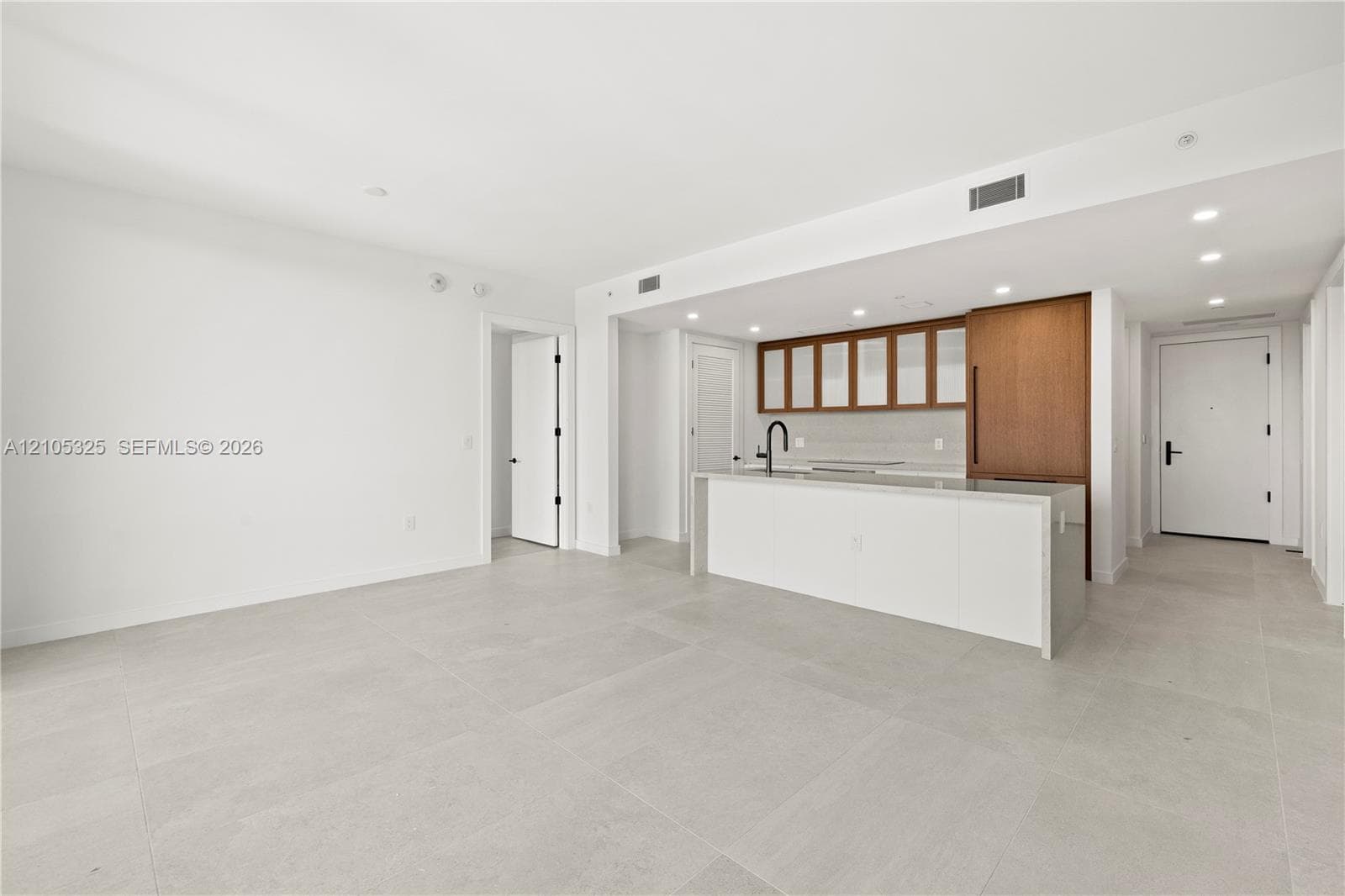 Open concept living space featuring light-toned tile flooring, recessed ceiling lighting, a kitchen island with integrated sink, wood-finish cabinetry, and a contemporary matte black faucet
