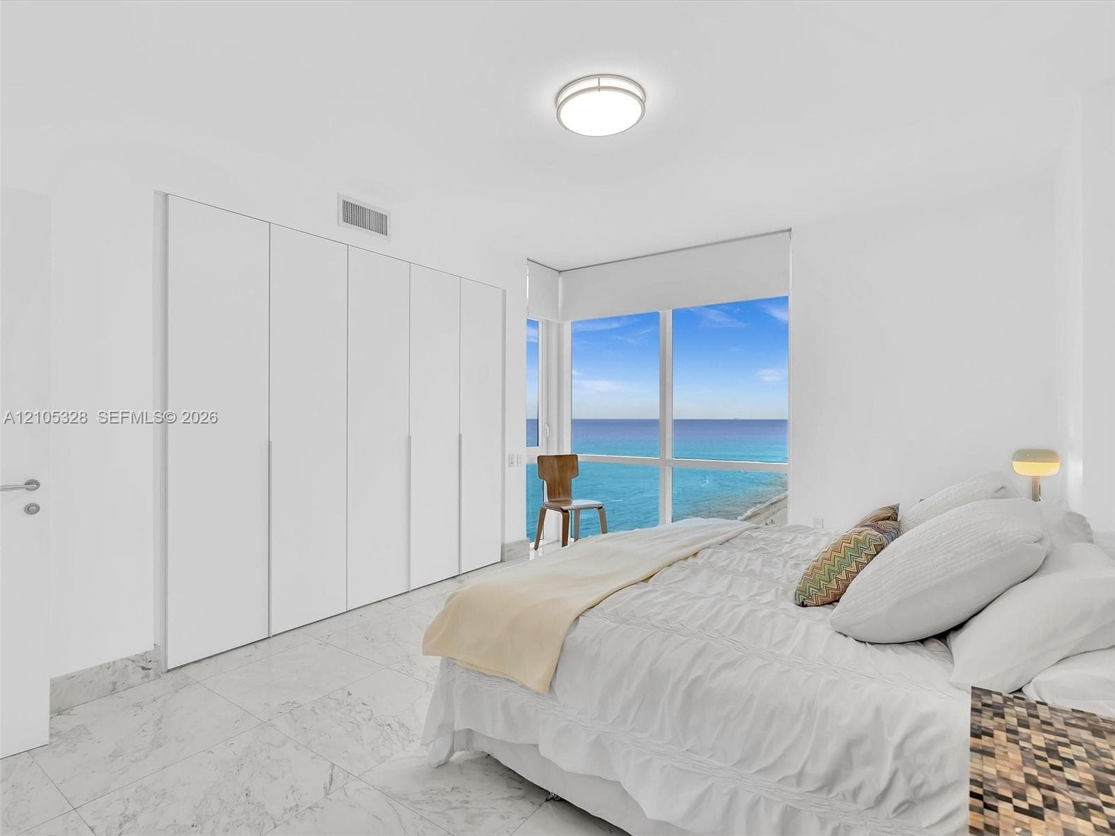 Bright and elegant bedroom featuring dramatic floor-to-ceiling windows, custom built-in wardrobe cabinetry, luxurious marble-look flooring, recessed ceiling lighting, and a sophisticated white color palette.