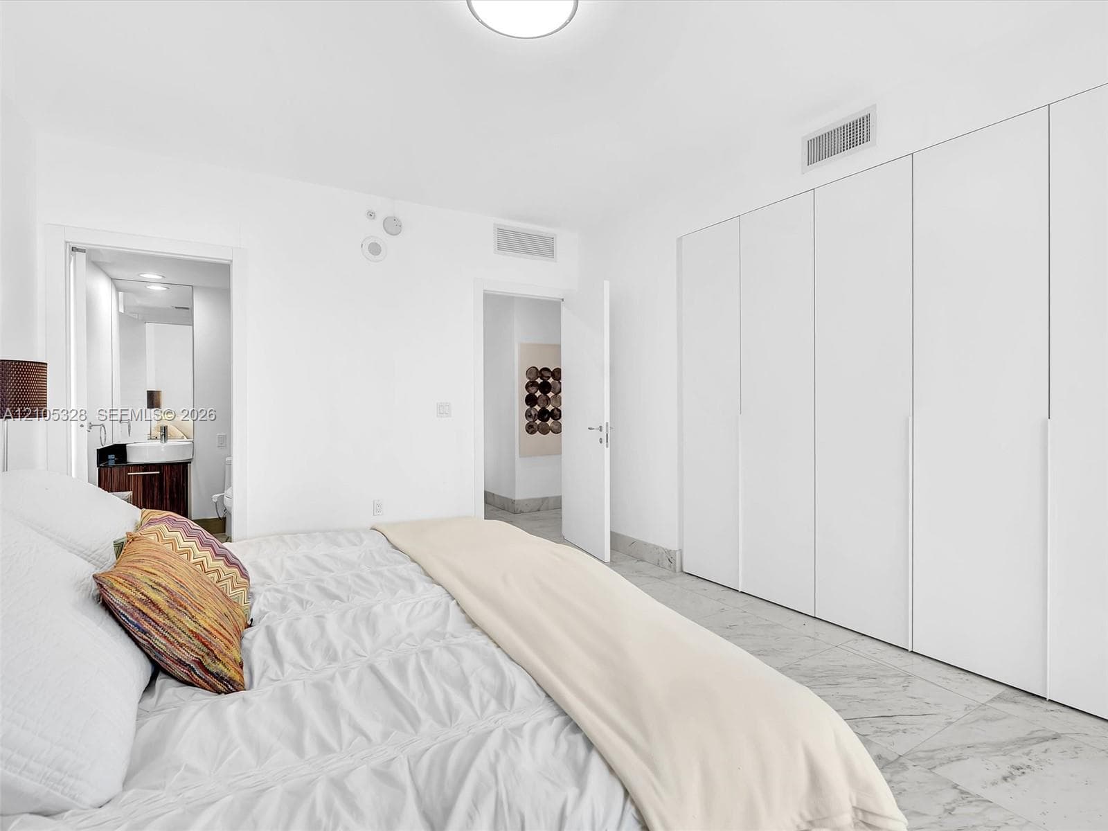 Bright and elegant bedroom featuring dramatic floor-to-ceiling windows, custom built-in wardrobe cabinetry, luxurious marble-look flooring, recessed ceiling lighting, and a sophisticated white color palette.