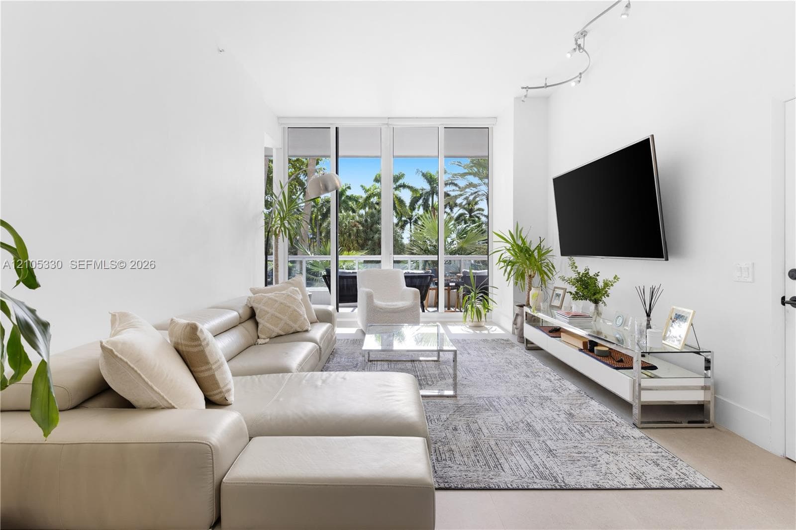 Bright, light-filled living space showcasing floor-to-ceiling glass doors opening to outdoor grounds, neutral wood-finish flooring, and a neutral-toned sectional arrangement