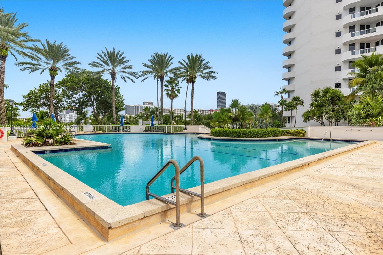 Expansive outdoor pool with integrated shallow section, surrounded by textured paving, lush landscaping, and mature palm trees