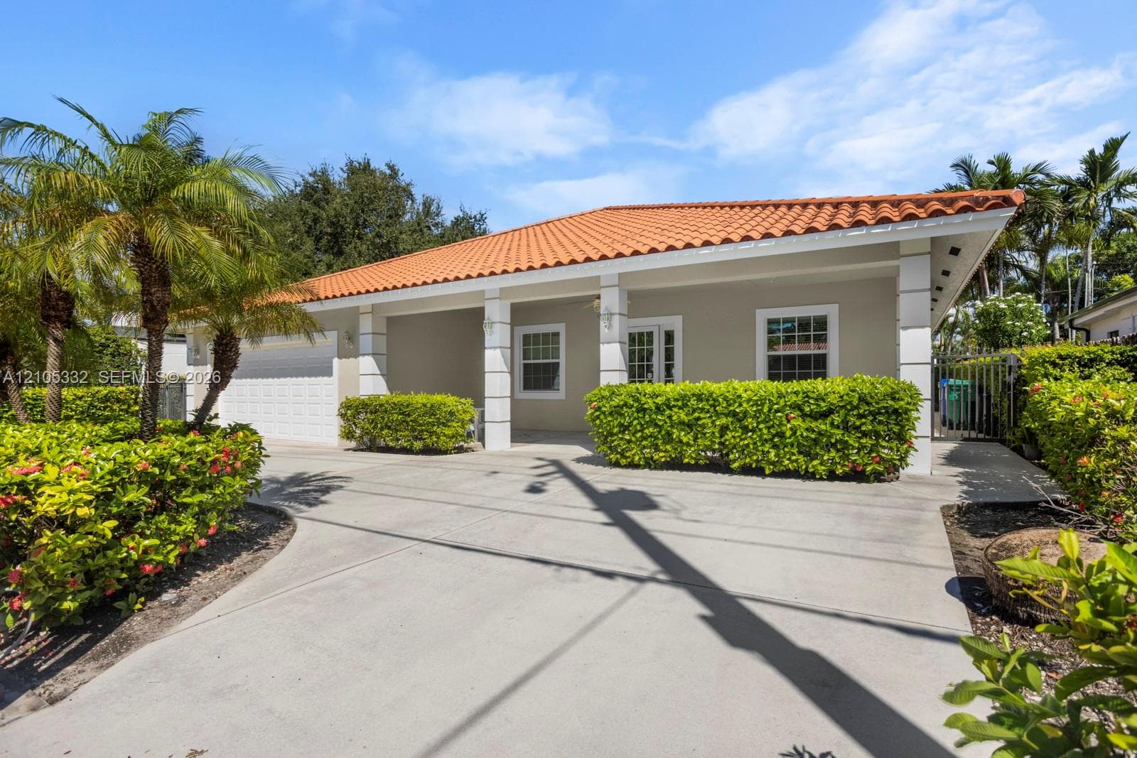 Concrete driveway leading to a residence with a terracotta tile roof, white exterior columns, and landscaping featuring palm trees and manicured hedges