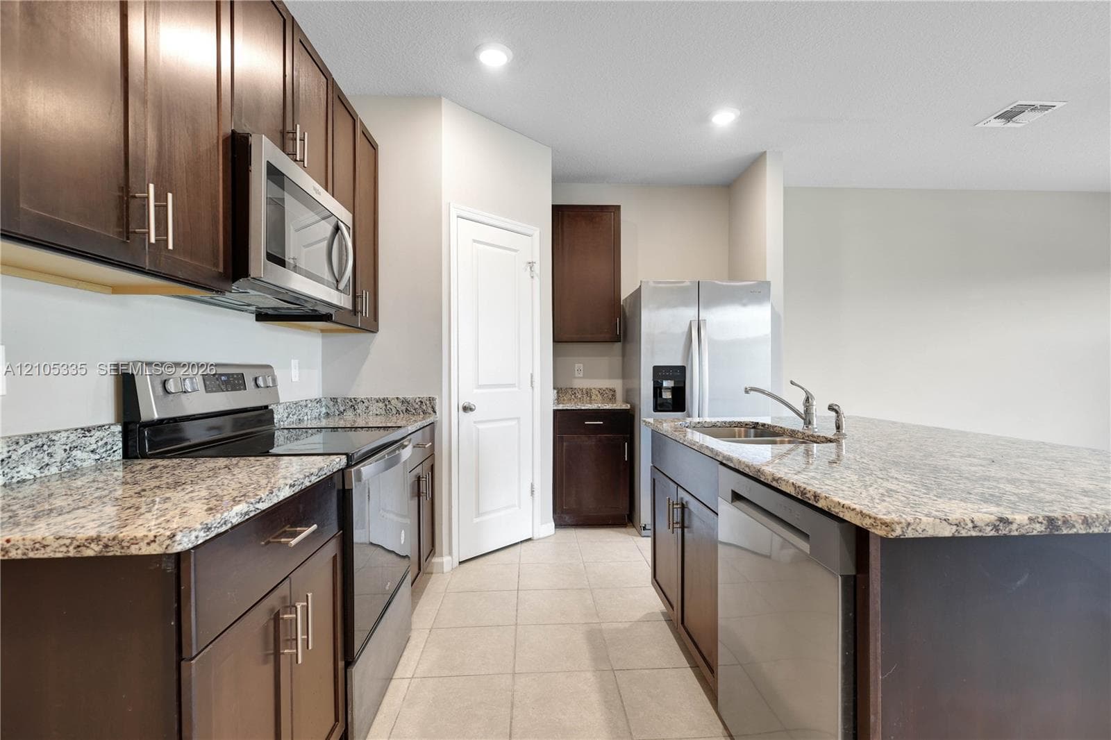 Kitchen featuring dark wood-finish cabinetry, light-toned granite countertops, stainless steel appliances, and tile flooring
