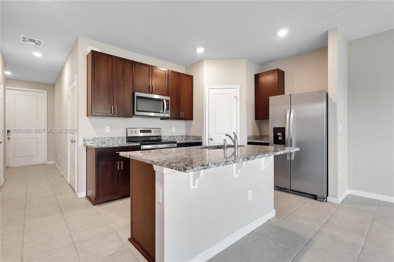Kitchen featuring an island with a built-in sink, stainless steel appliances, granite countertops, dark wood-finish cabinetry, and recessed lighting