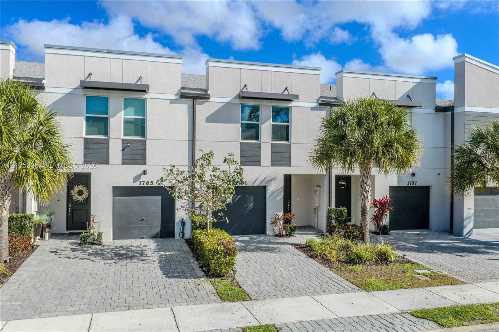 Modern townhome exterior with stucco finish, dark gray garage doors, paver driveways, and mature landscaping