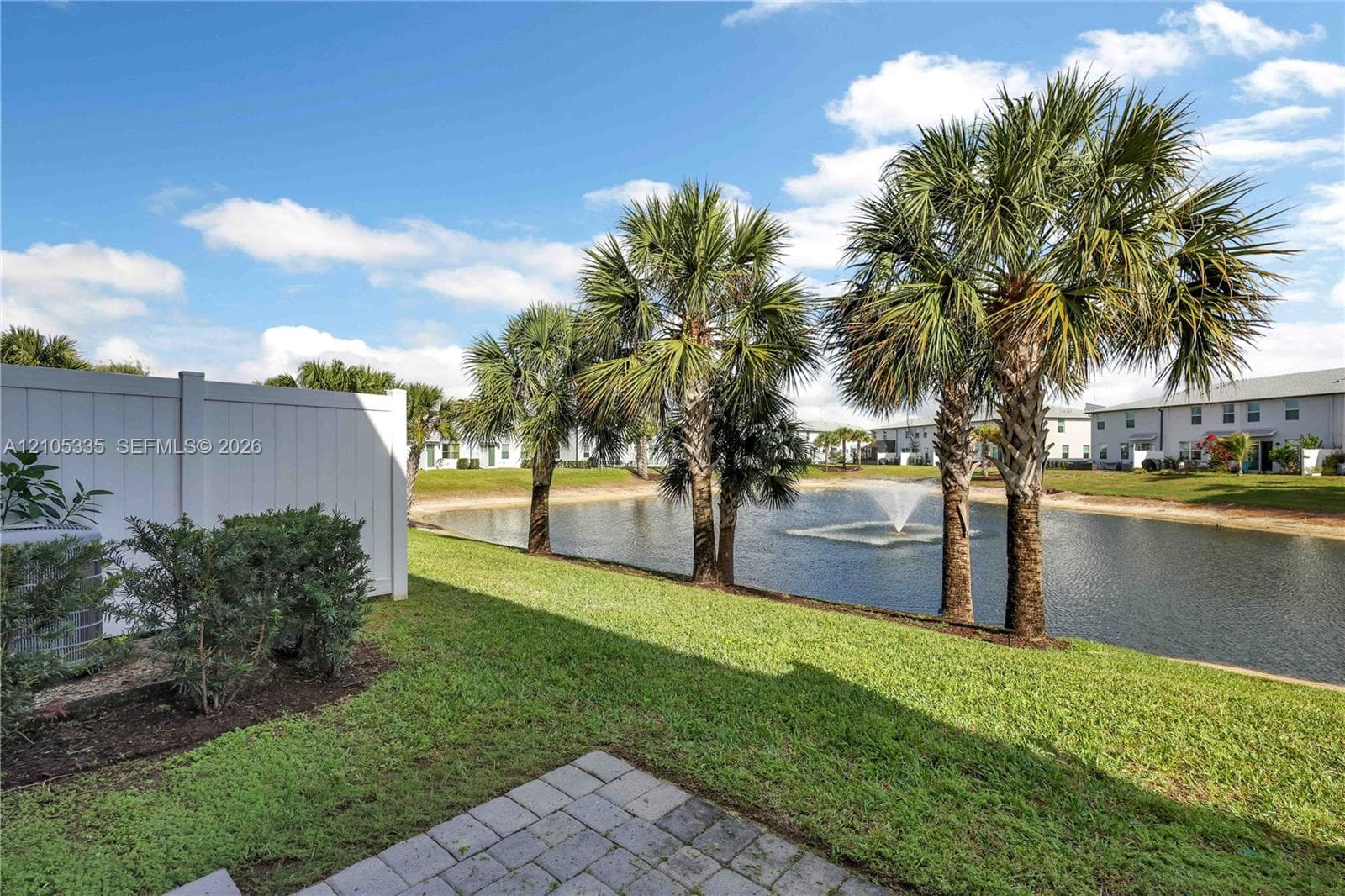 Paver patio with a white privacy fence, mature landscaping, and waterfront access featuring a fountain