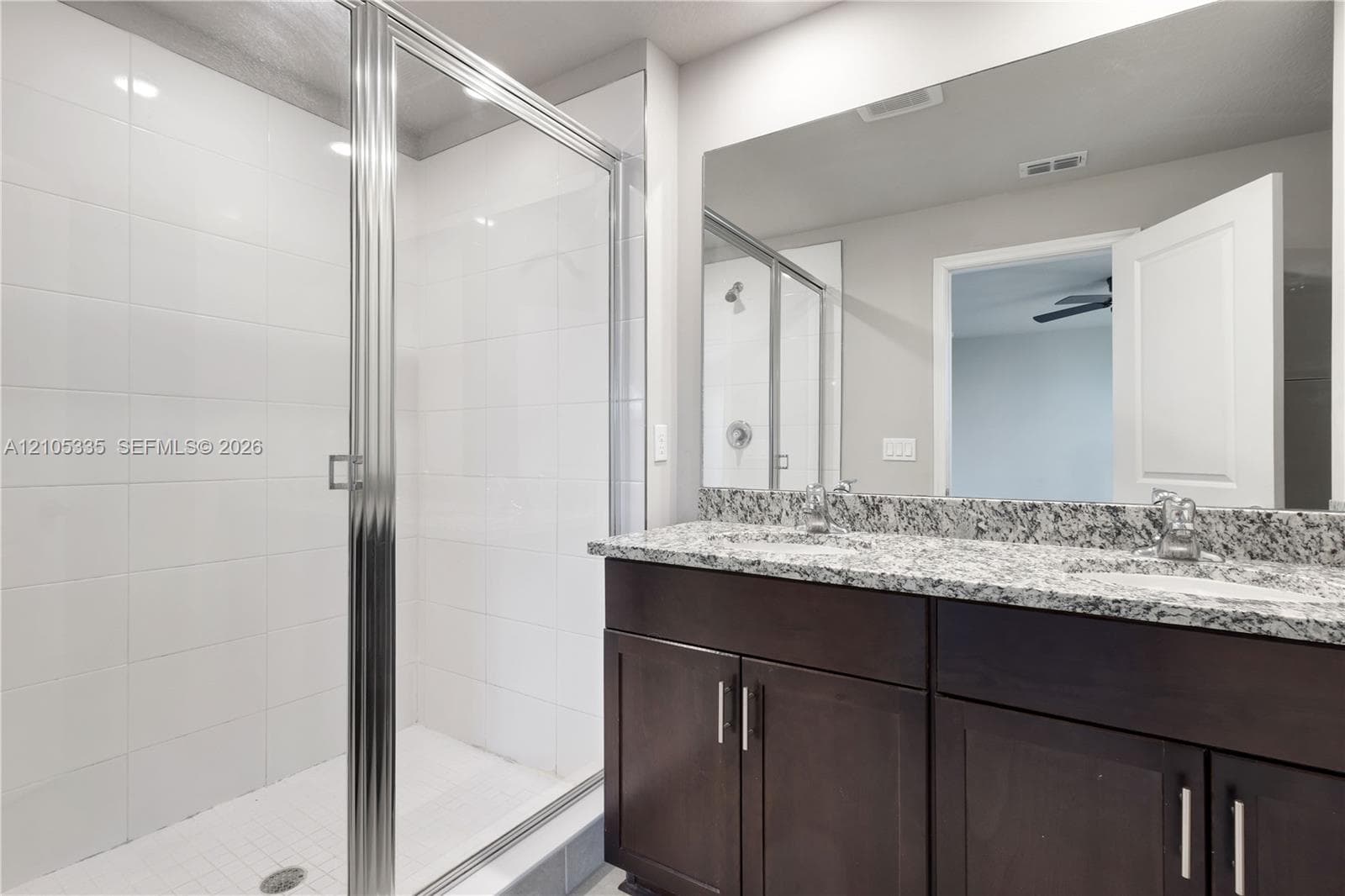 Glass-enclosed shower with white tiled walls, dual vanity with granite countertop, and dark wood-finish cabinetry