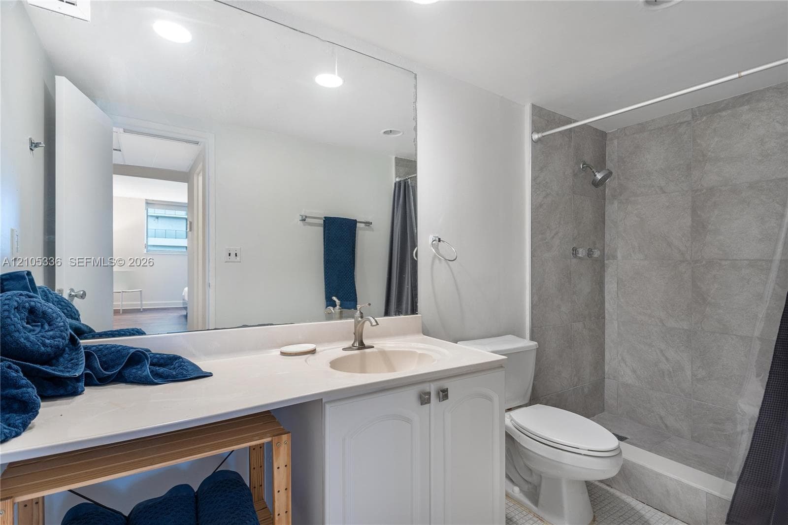Bathroom featuring a vanity with a white countertop and integrated sink, a full-width wall mirror, and a shower enclosure with floor-to-ceiling stone-finish tile surround