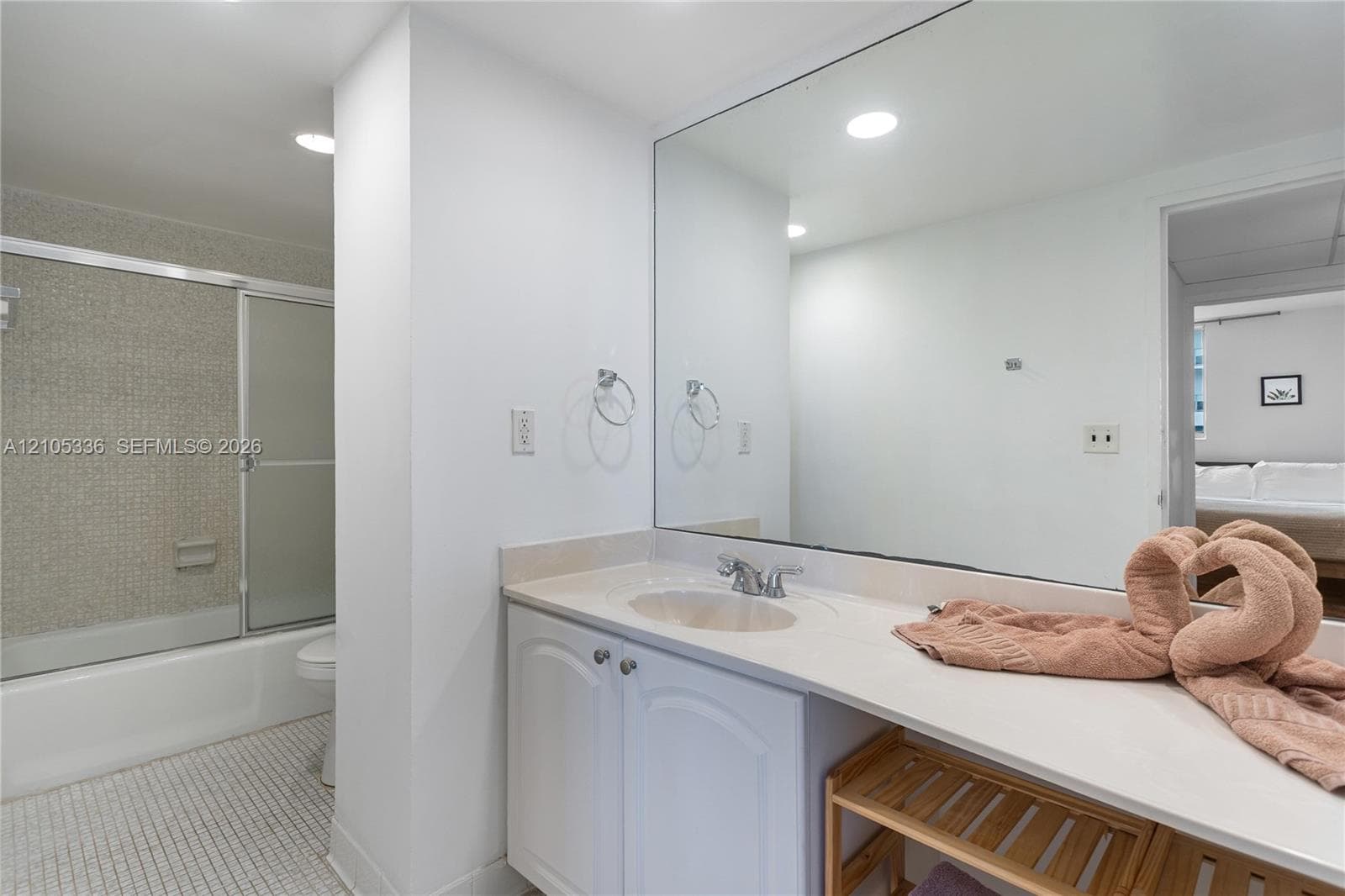 Bathroom vanity with white cabinetry, solid-surface countertop, integrated basin, large wall-mounted mirror, and tub-shower combination featuring textured wall tile and mosaic floor tile