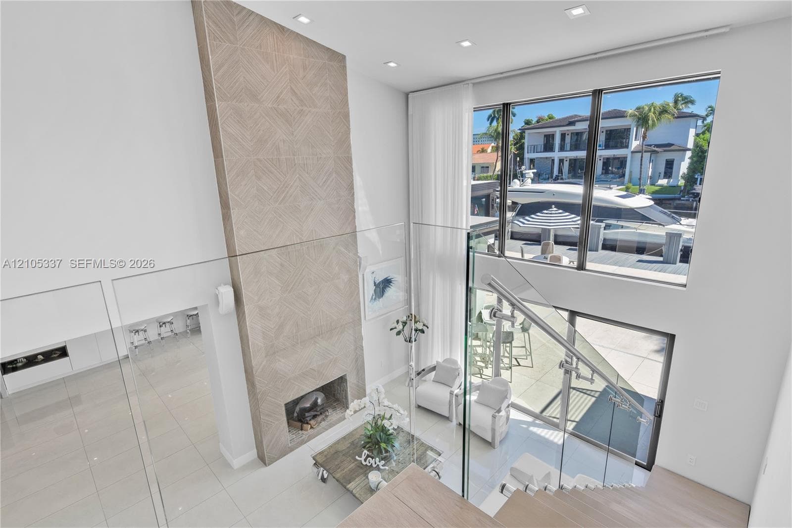 Double-height living area featuring floor-to-ceiling windows, full-height decorative tile fireplace surround, recessed lighting, and glass panel stair railing