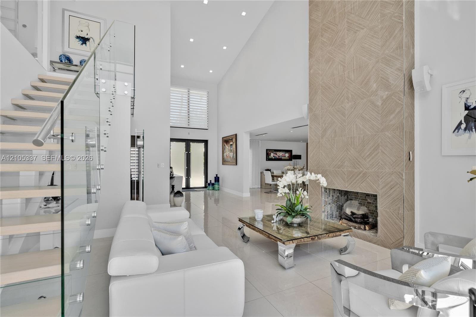 Double-height ceilings, floating staircase with glass panel railing, floor-to-ceiling tiled fireplace surround, and recessed lighting