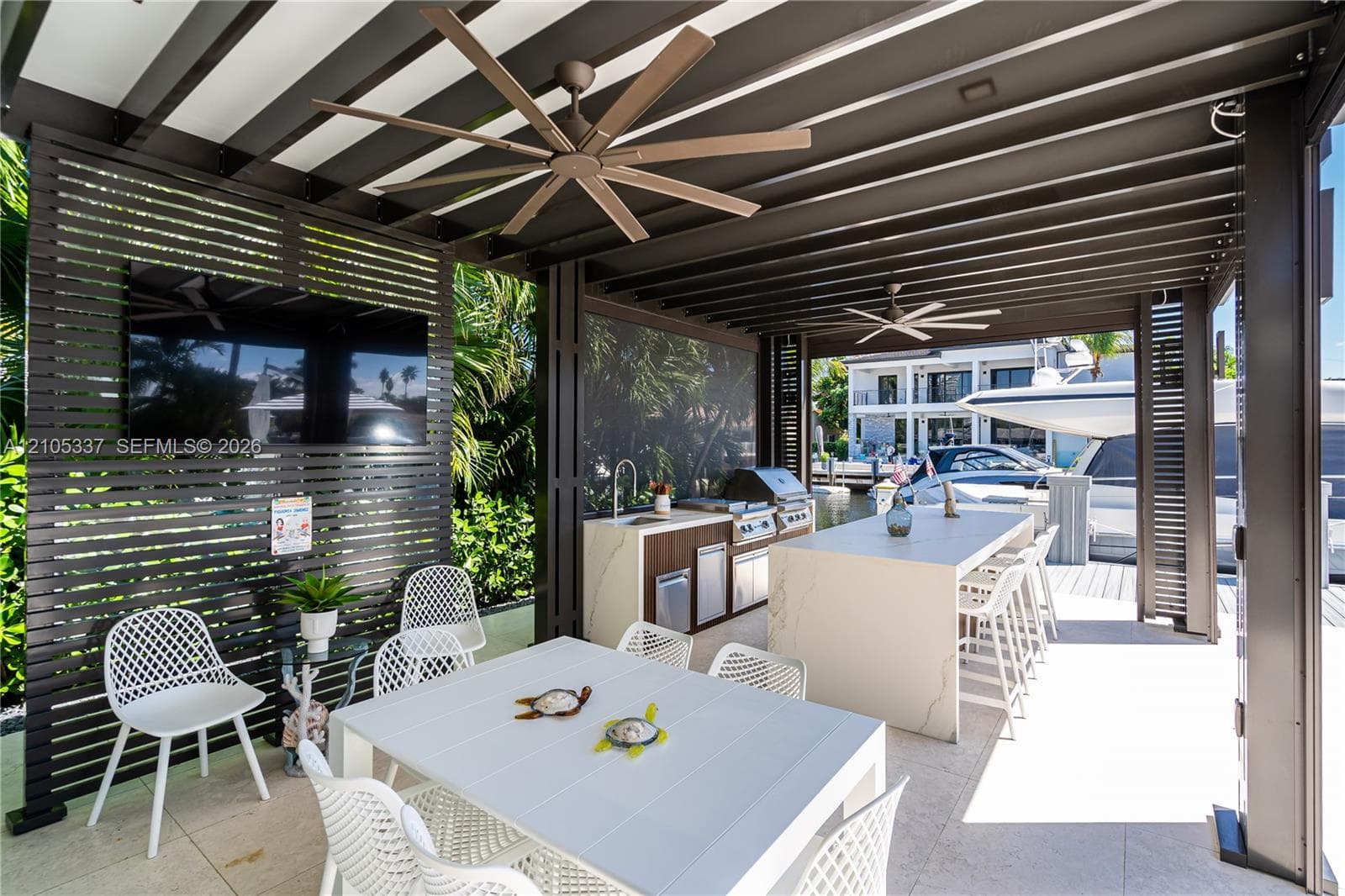 Covered outdoor kitchen featuring a stone-topped island with integrated seating, built-in stainless steel appliances, slatted wood-finish privacy screens, and dual ceiling fans