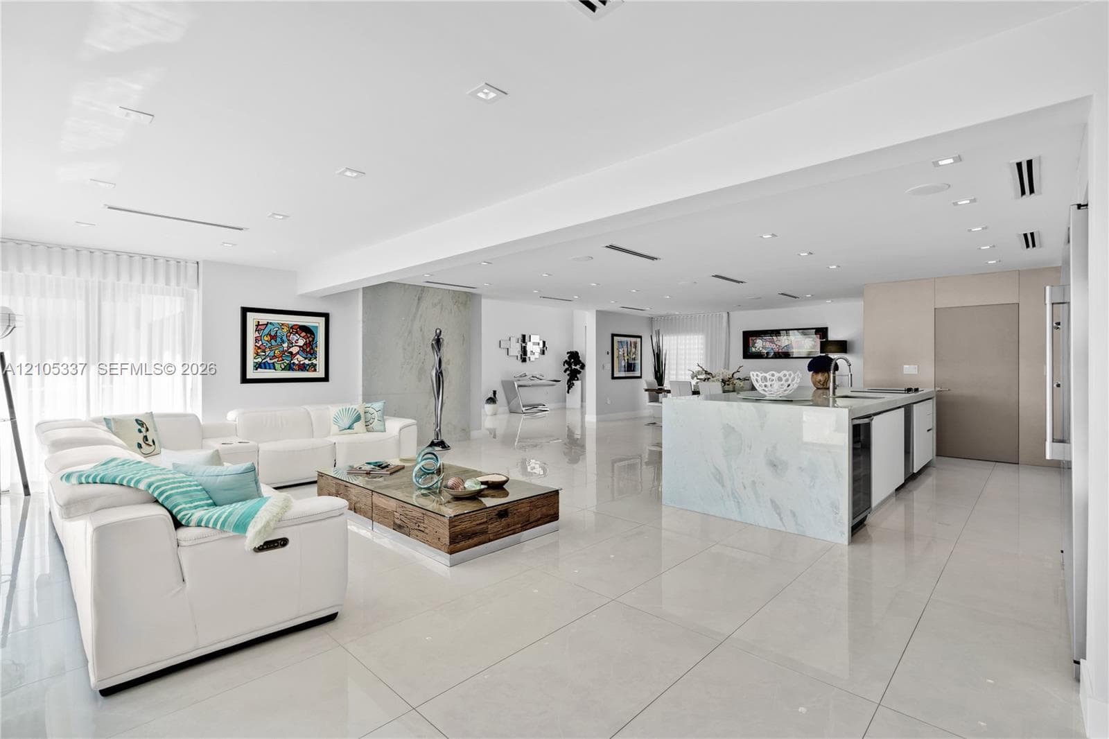 Open-concept interior featuring large-format tile flooring, recessed ceiling lighting, a waterfall-edge kitchen island, and a full-height stone fireplace surround