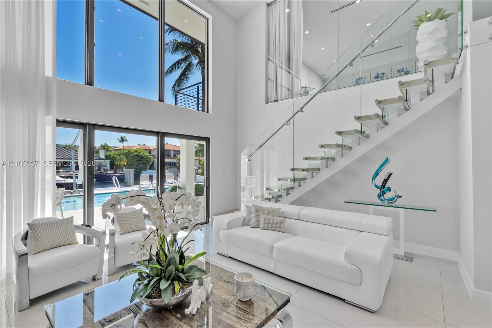 Double-height ceilings featuring floor-to-ceiling windows, open-riser staircase with glass balustrades, and sliding glass doors leading to an exterior pool area