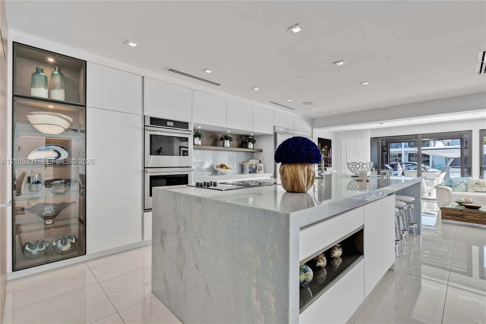 Modern kitchen featuring floor-to-ceiling glass-front cabinetry, a waterfall-edge stone island, built-in dual wall ovens, recessed ceiling lighting, and tile flooring
