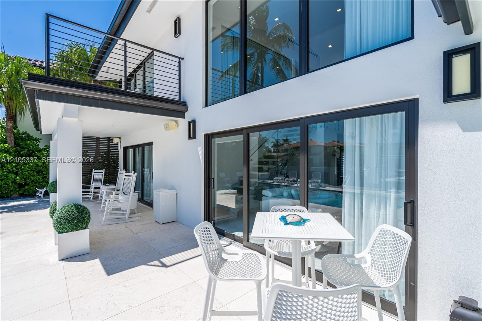 Two-story exterior with white stucco finish, expansive floor-to-ceiling glass sliding doors, upper-level balcony with black metal railing, and covered ground-level patio featuring light-toned stone tile flooring