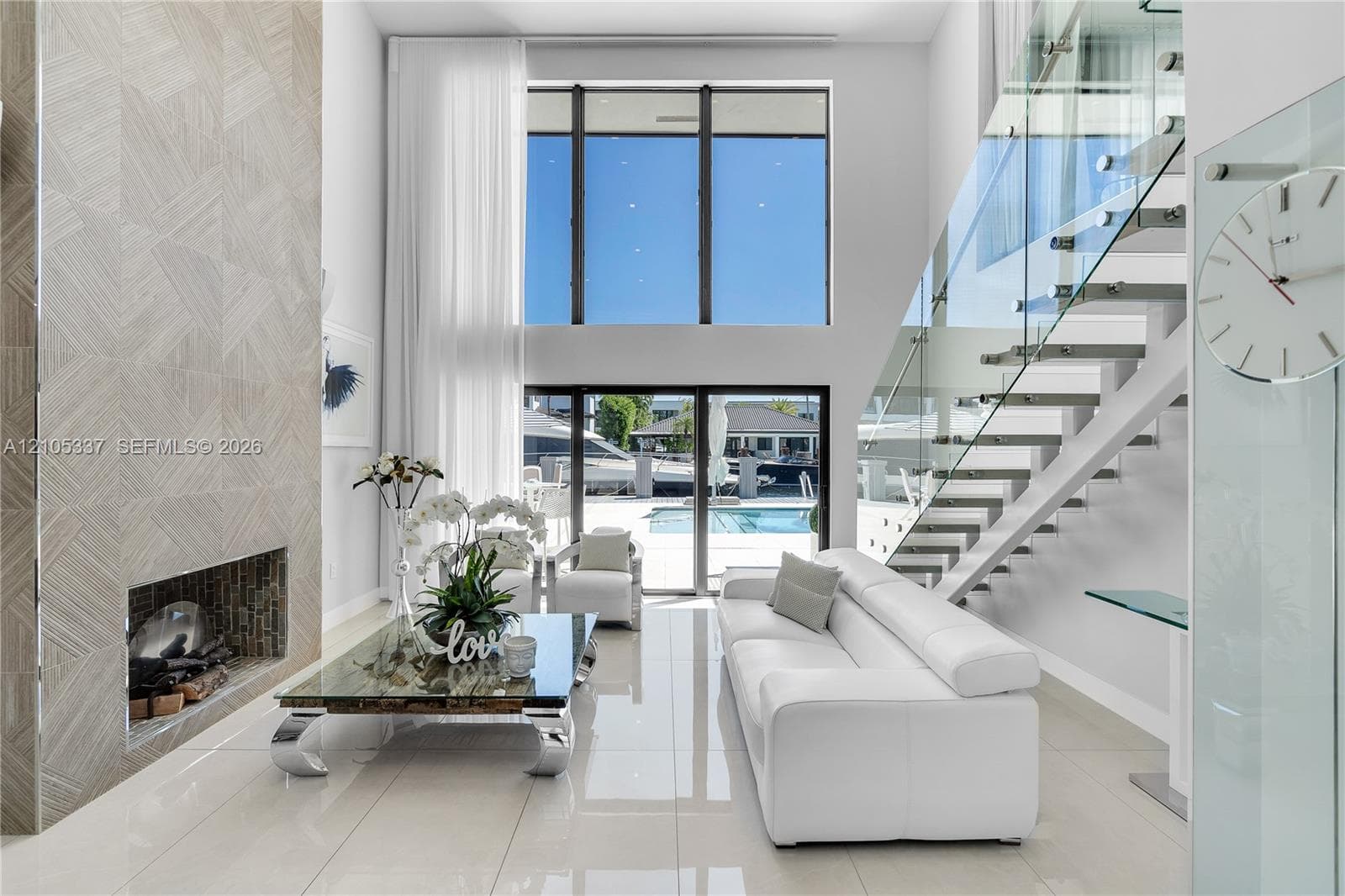 Double-height living area featuring floor-to-ceiling windows, a floor-to-ceiling textured tile fireplace, polished tile flooring, and a staircase with glass railings and metal treads