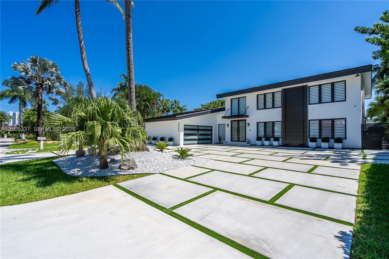 Modern two-story exterior featuring white stucco finish, geometric concrete paver driveway with turf accents, multi-pane horizontal glass garage door, and metal-trimmed window groupings