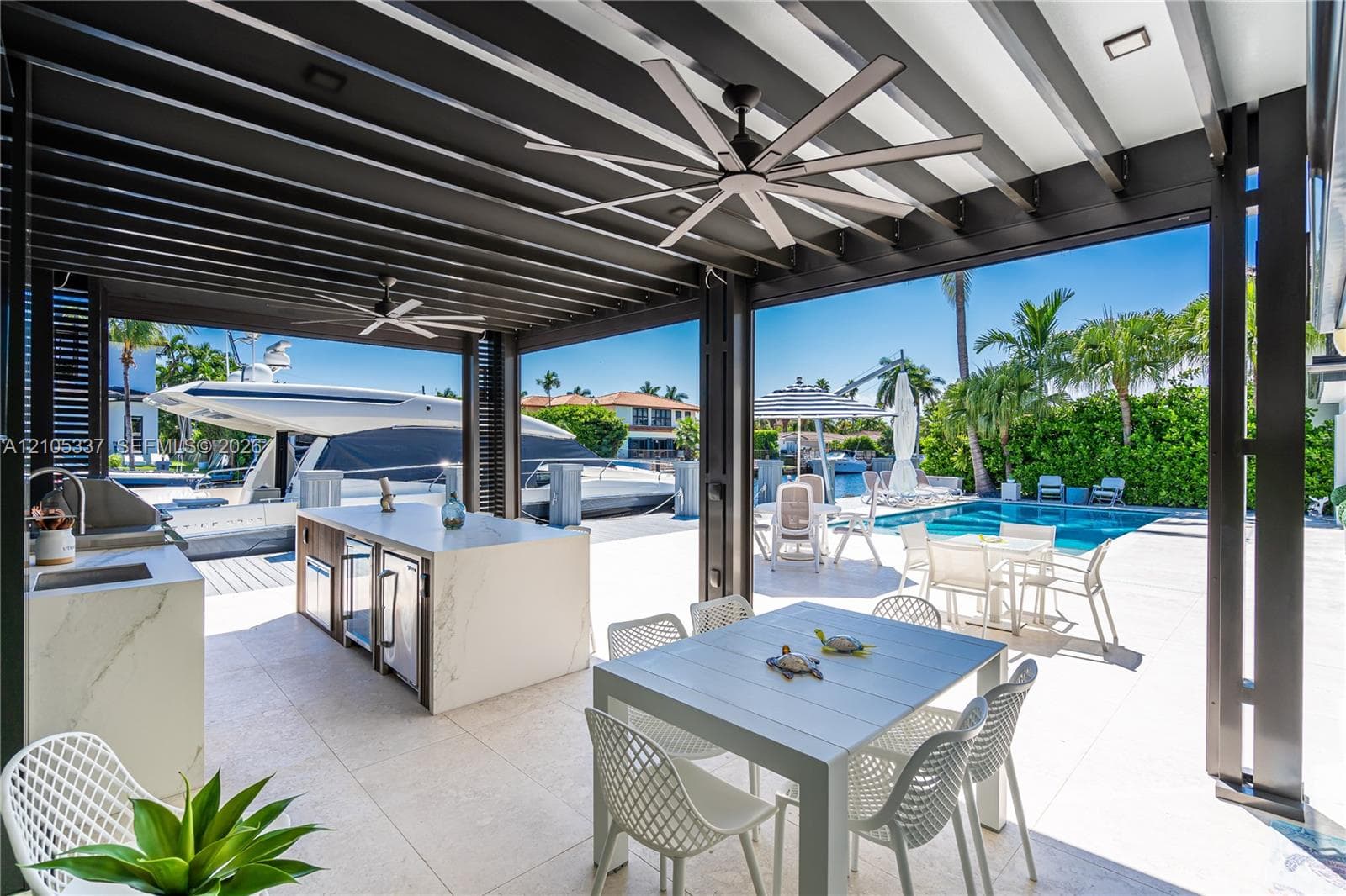 Covered outdoor kitchen featuring a stone-clad island, stainless steel built-in grill, and stone-finish flooring beneath a slatted pergola with multi-blade ceiling fans