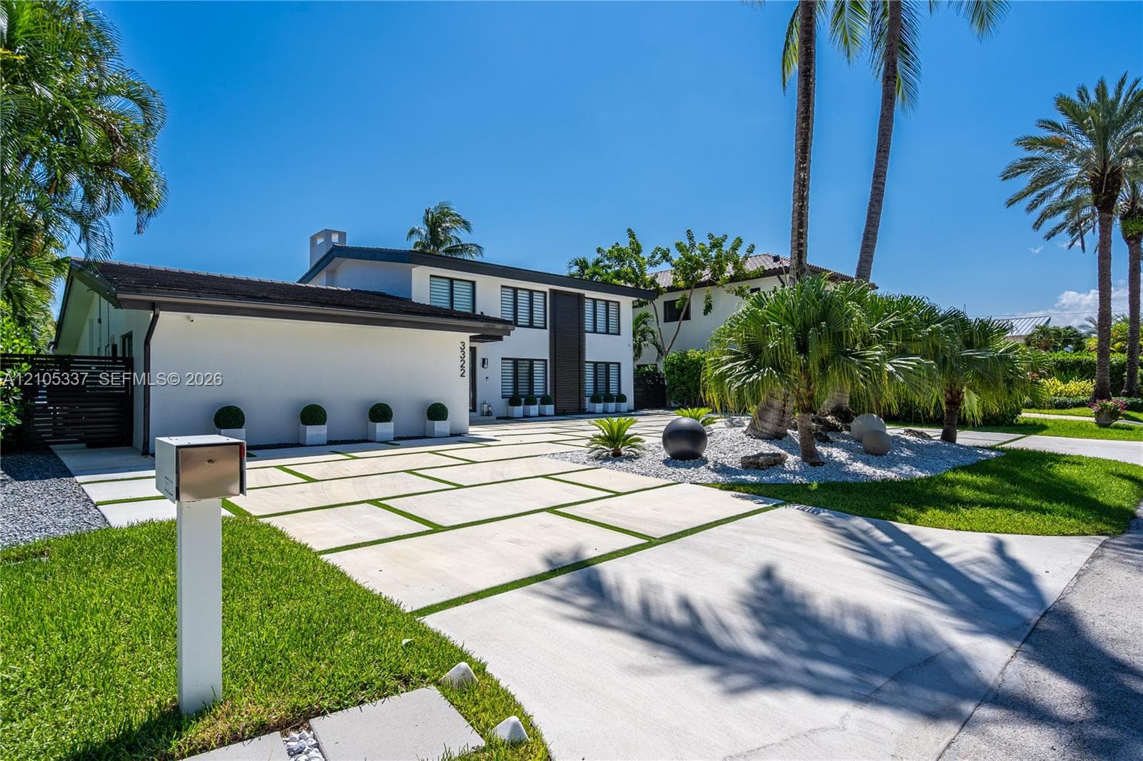 Modern two-story exterior featuring smooth stucco siding, large paneled windows, a grid-pattern concrete driveway with integrated turf, and landscaped grounds with mature palm trees