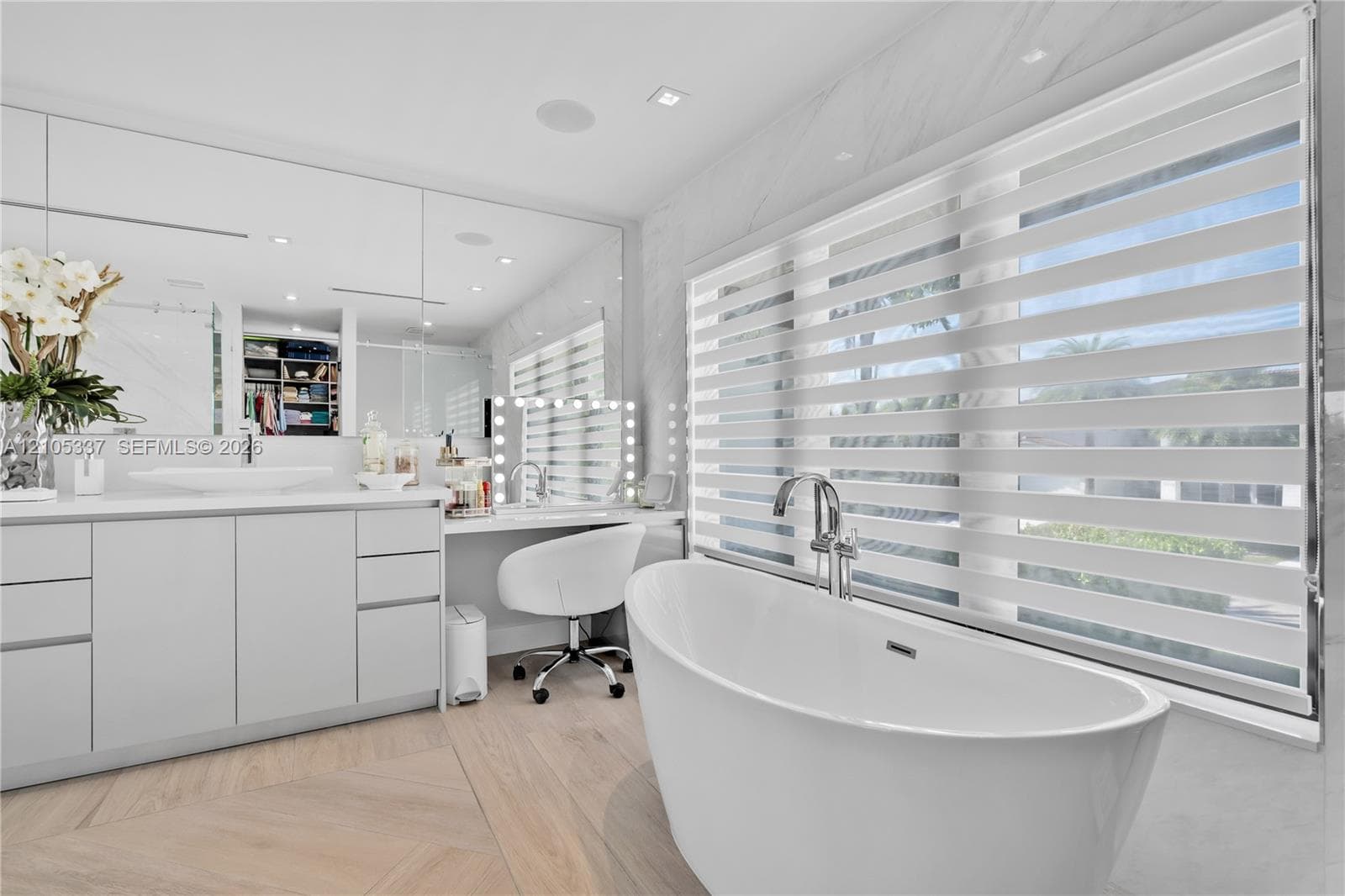 Freestanding soaking tub with a chrome floor-mounted faucet, custom vanity with integrated sink and stone-finish countertop, full-wall mirror, and wood-finish flooring in a herringbone pattern