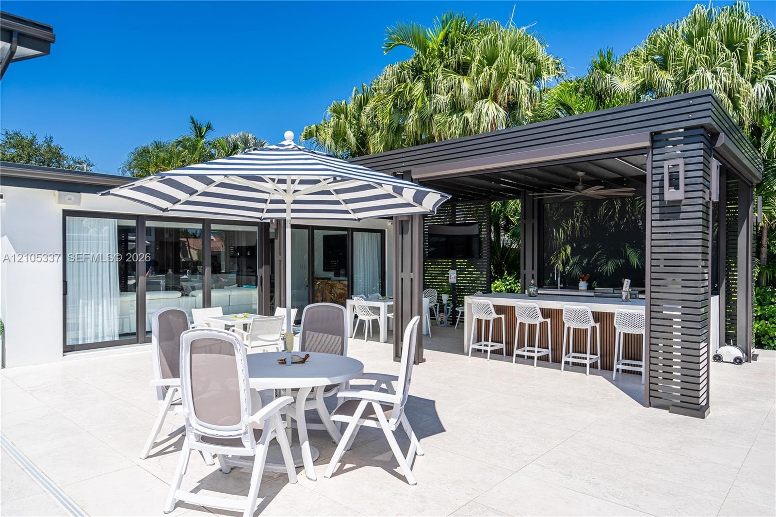 Outdoor patio featuring large-format light-toned tiling, a covered cabana with a built-in stone-top bar, recessed ceiling fan, and sliding glass doors