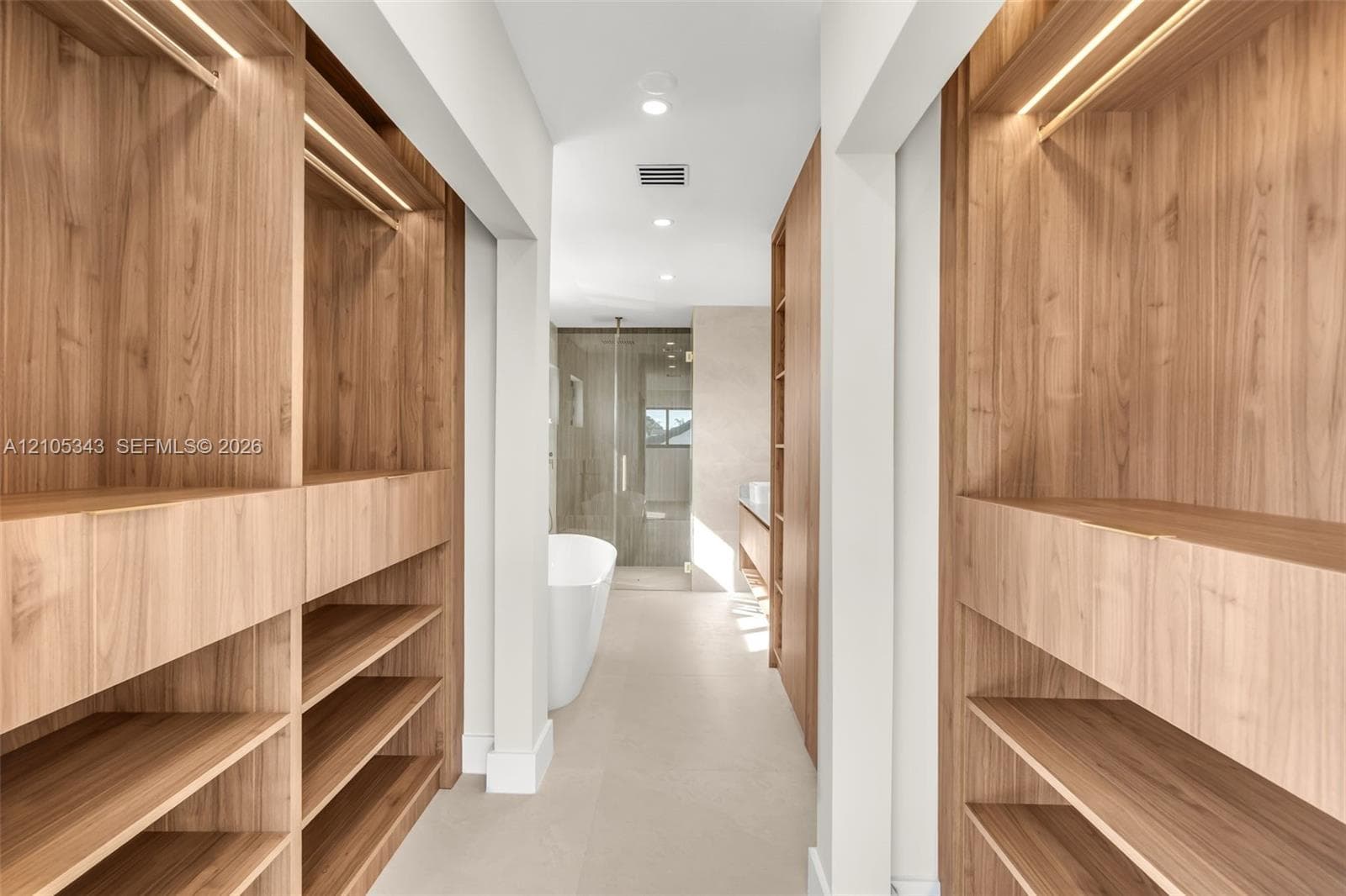 Custom wood-finish cabinetry with integrated lighting, open shelving, freestanding soaking tub, glass-enclosed shower, and large-format tile flooring